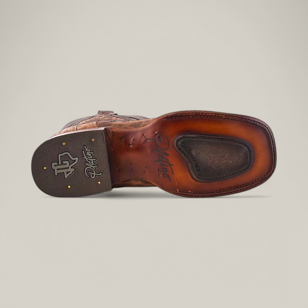 The image showcases the sole of the Exotic Carunga - Diamond cowboy boot, featuring smooth brown leather and intricate stitching. The heel has a logo with decorative elements, accented by two gold rivets on each side, set against a plain light grey background.