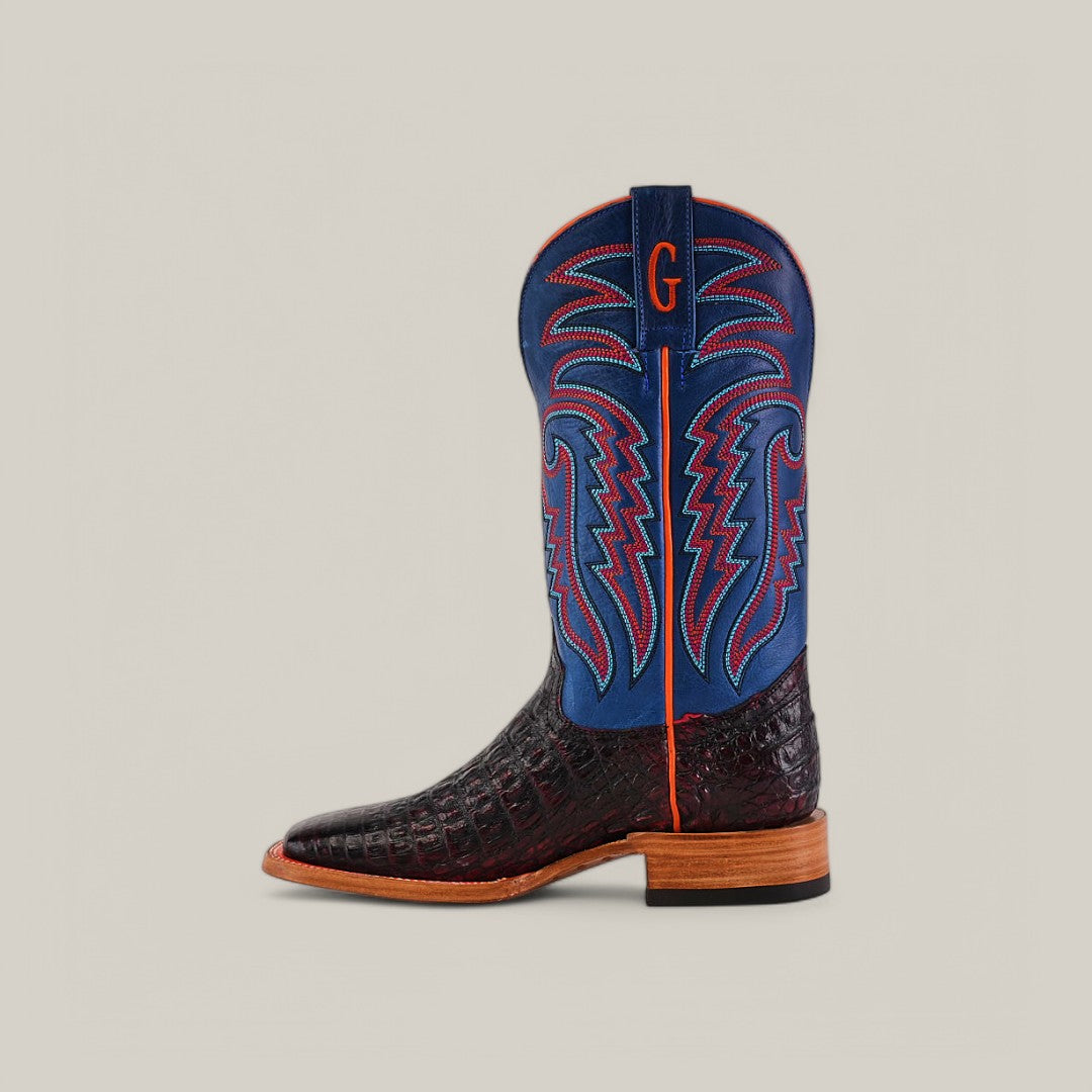 The Exotic Caiman Belly - Black Cherry cowboy boot features a dark brown crocodile-patterned lower, a blue upper with red and blue stitching, a wooden sole, and a handcrafted red G on the blue section near the top, all set against a neutral background.