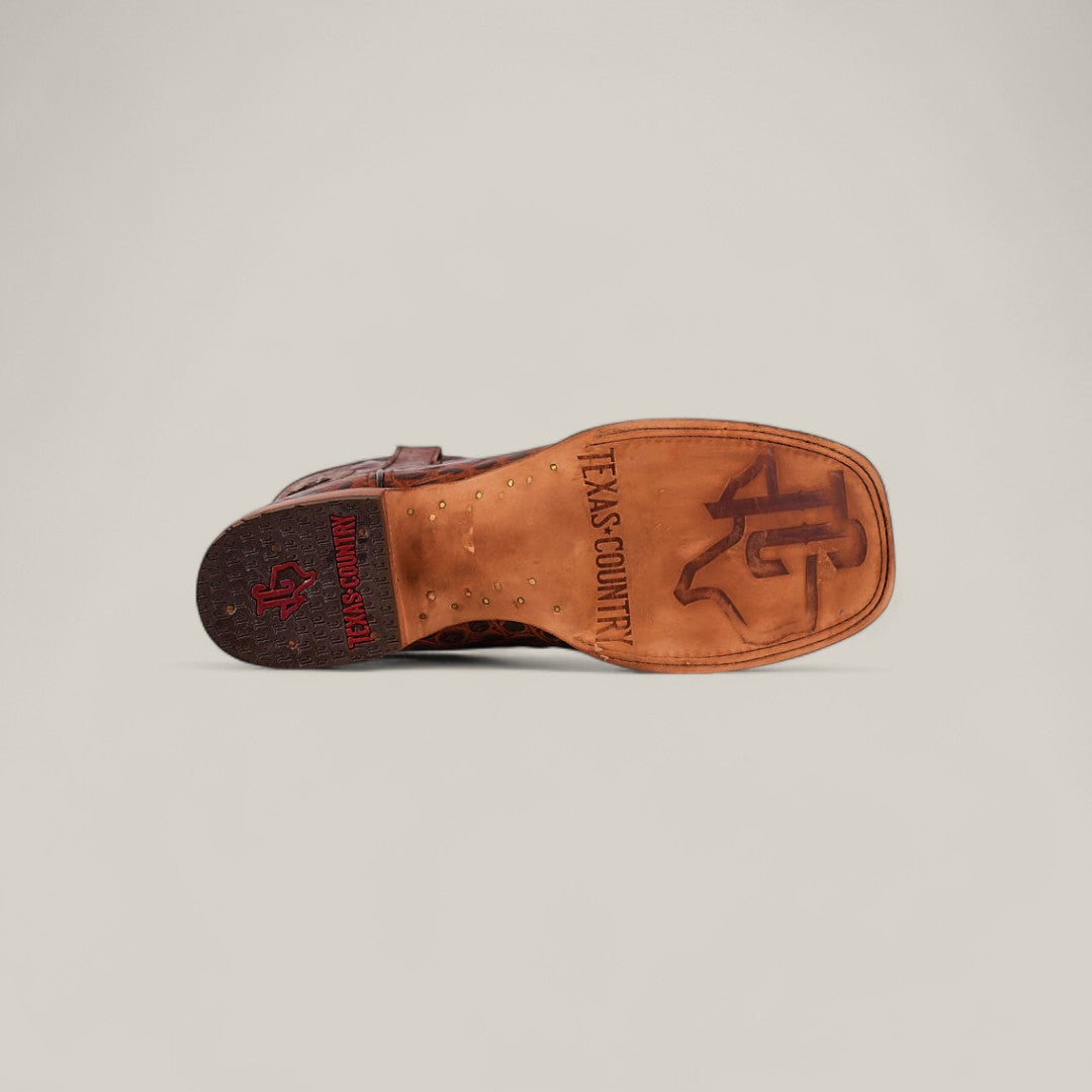 The Jonh - Cogñac - Square Toe boot is displayed sole-up, revealing the Texas A&M logo and “TEXAS COUNTRY” engraved on the sole. The heel shows a red TEXAS COUNTRY logo and a small Texas outline.