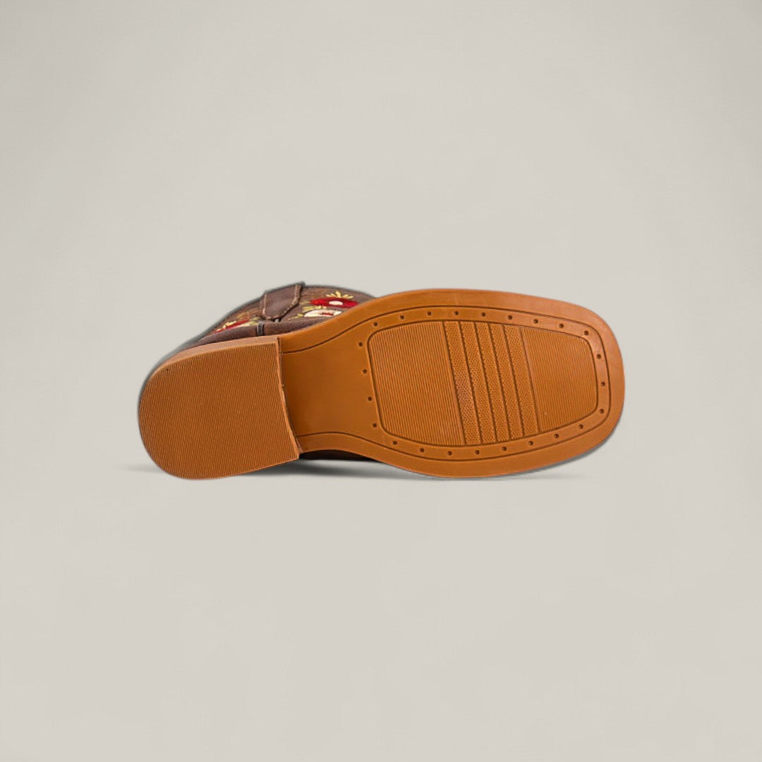 The Karo Brown - Square Toe - Kids Boots is shown lying on its side, featuring embroidered floral designs on brown leather and a flat, light brown rubber sole with textured grip patterns, blending comfort and elegance against a plain background.