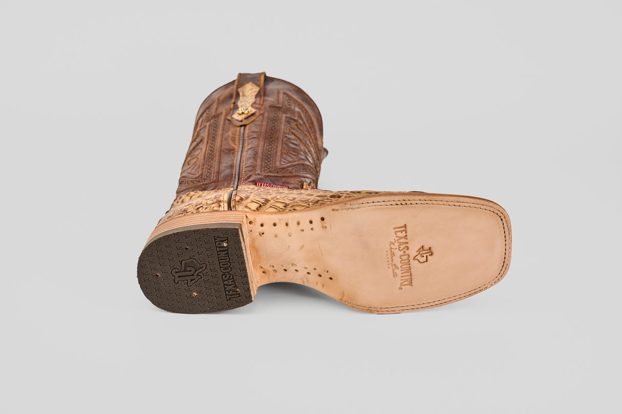 A single Exotic Caiman Horn Back - Antique square toe boot lies on its side, displaying detailed stitching on the upper and a light tan sole with Texas Country branding. This cowboy boot also features a rubber heel cap.