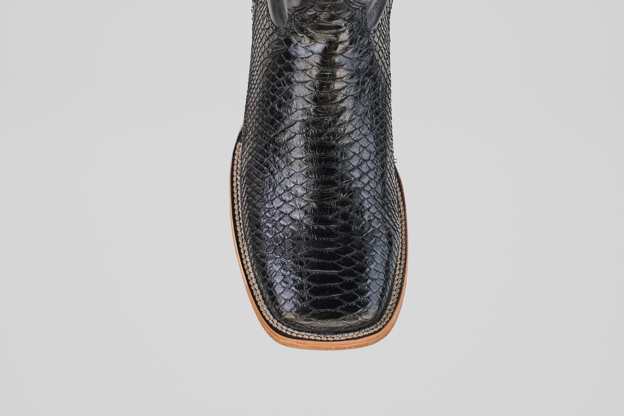 A close-up top view of the Tanner - Black - Square Toe, a black cowboy boot with a shiny, textured Python Negro snakeskin pattern and a light brown and white stitched sole.