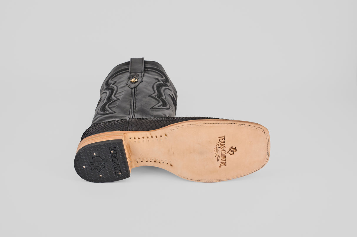 A Tanner - Matte Black - Square Toe cowboy boot lies on its side, displaying the textured matte black upper and the tan sole with brand and stitching details visible underneath.