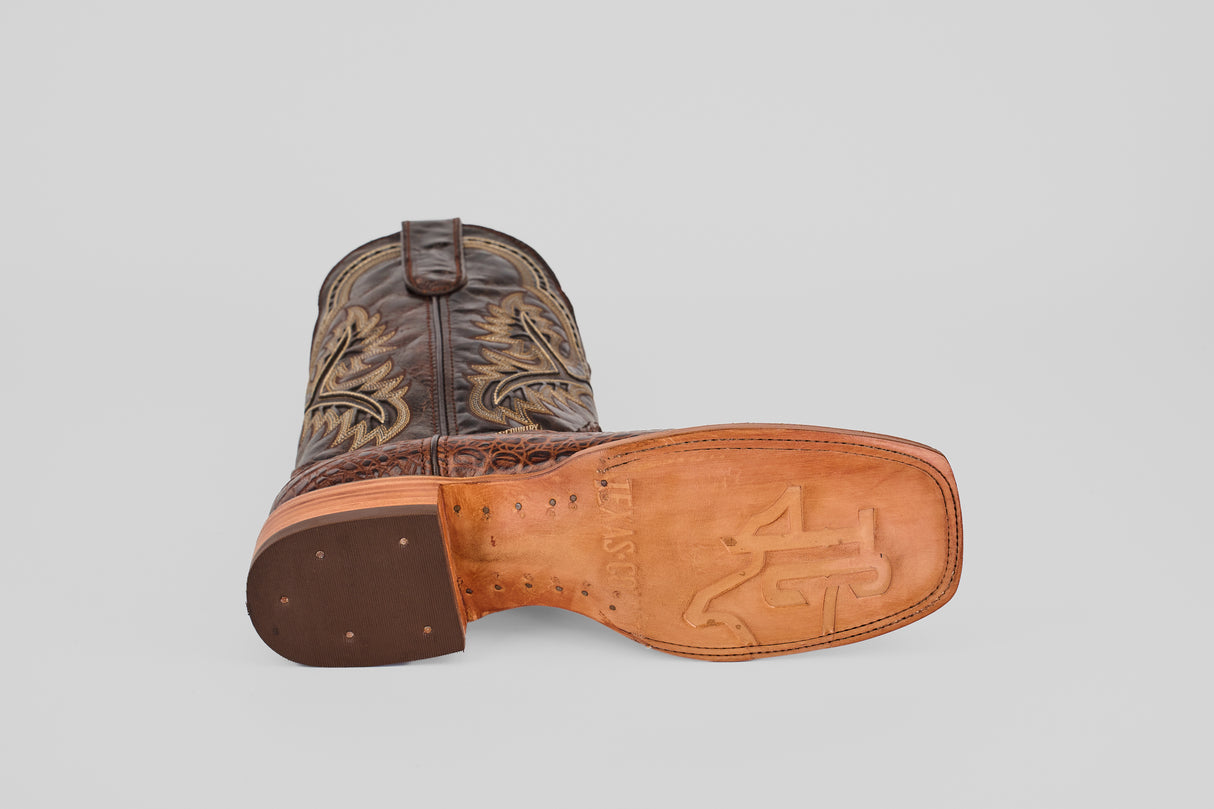 The Caiman Belly Choco - Embossed Leather - Square Toe (E423) cowboy boot lies on its side, showcasing decorative stitching, a light tan sole with "A" and "1" logos, and small metal studs near the heel for classic craftsmanship.