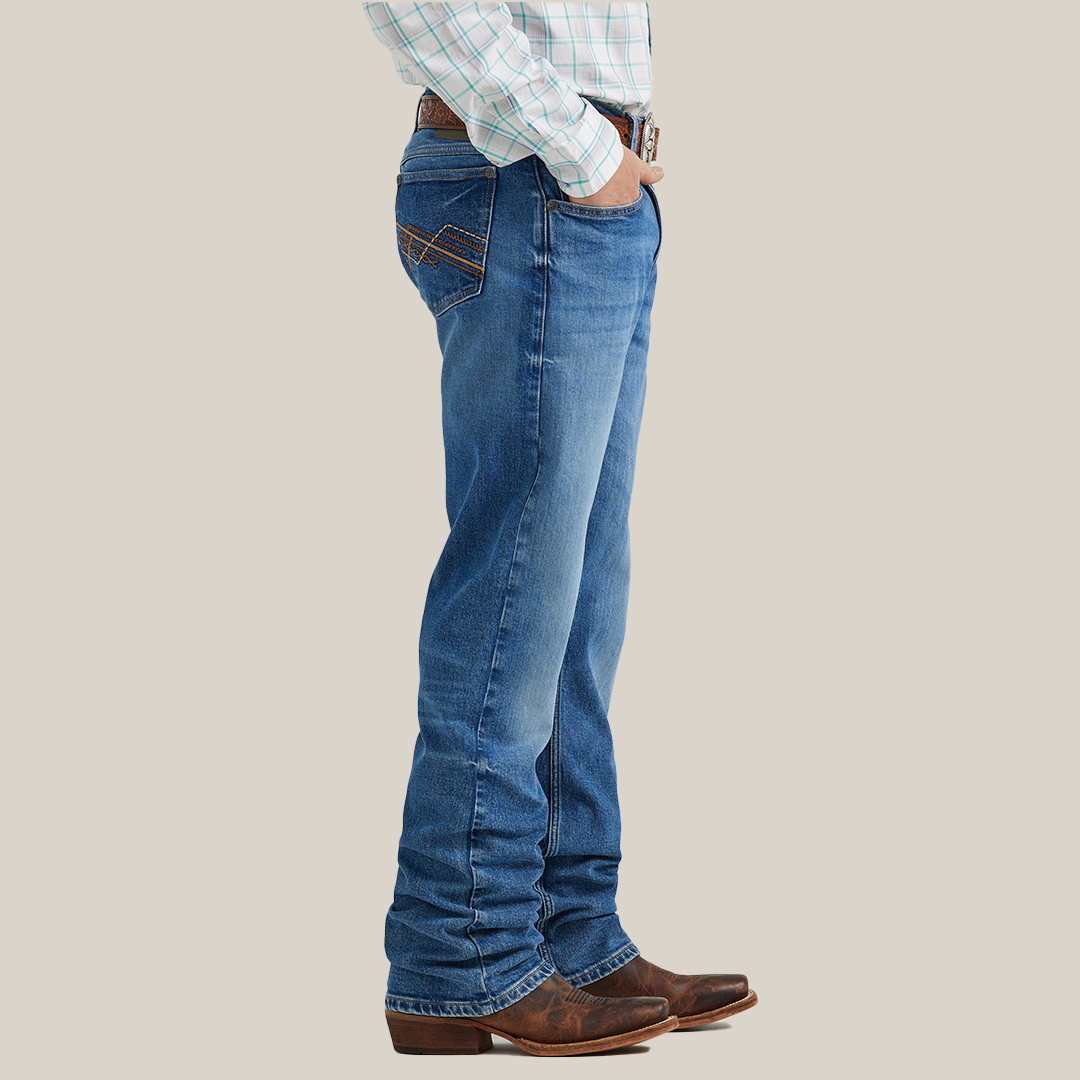 A person wearing Wrangler 20x Mens Medium Wash Backwater Vintage Bootcut Stretch Denim Jeans, a plaid shirt, and brown cowboy boots stands in profile with hands in pockets against a neutral background.