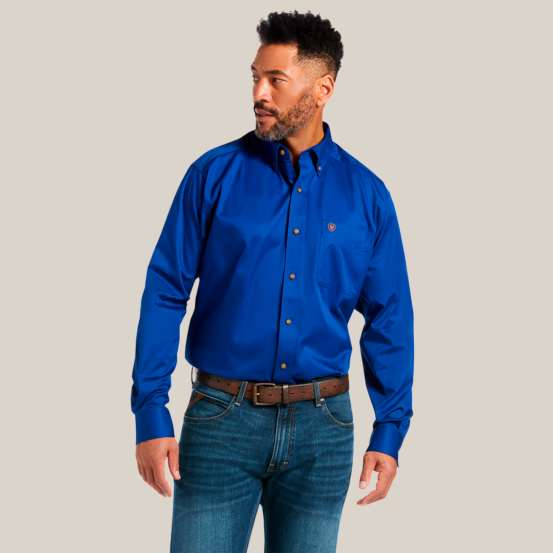 A man with short curly hair and a beard wearing a Solid Twill Classic Fit Shirt Blue - 10006660 and blue jeans with a brown belt stands looking to his right. The 100% Cotton Twill shirt showcases a small logo on the chest.