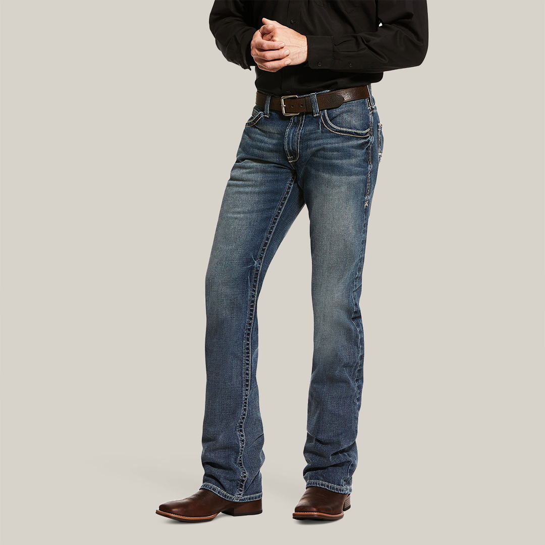 A person wearing a black long-sleeve shirt and M5 Slim Stretch Adkins Stackable Straight Leg Jean in lennox, accented with a brown belt and cowboy boots, stands against a plain white background with their hands clasped in front.