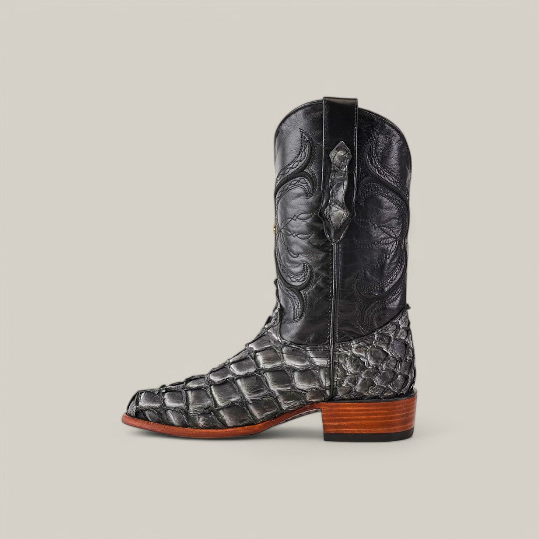 The Exotic Pirarucu Fish boot features a black textured design crafted from luxurious fish leather, with intricately stitched uppers and a smooth, square toe. It boasts a wooden heel, side pull strap, and is elegantly displayed against a plain beige background.