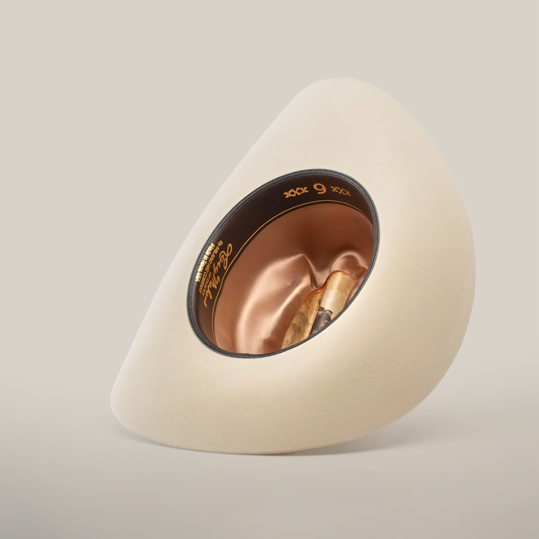 The 6X Real Belly is a beige, wide-brimmed fur felt hat, displayed upside down to reveal its satin-lined interior. The inside band features gold lettering and the number 9, all elegantly set against a light, plain background.