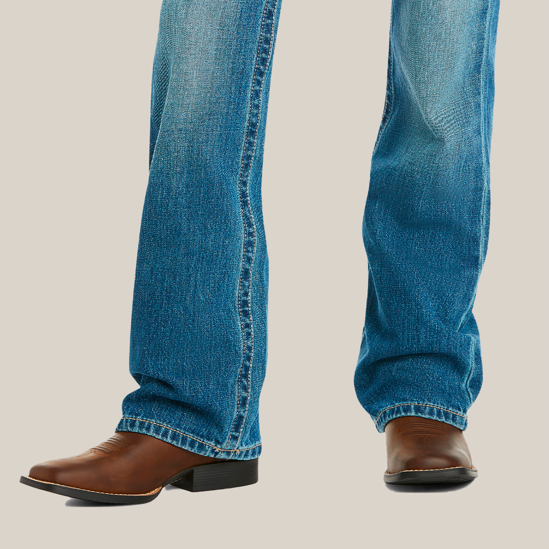A young boy stands with legs slightly apart, wearing Ariats medium wash boot cut jeans with a faded look and brown leather cowboy boots featuring a pointed toe and block heel, set against a white background.