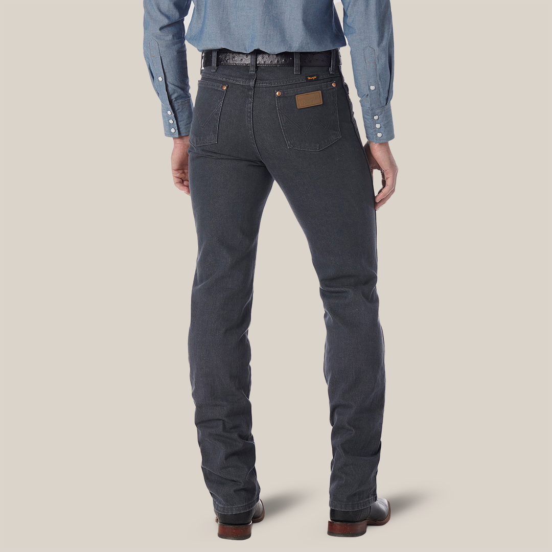 Rear view of a person in dark gray Wrangler Cowboy Cut Slim Fit Jeans - 0936CHG, a blue denim shirt with rolled-up cuffs, and brown shoes. A leather patch graces the back pocket. Set against a plain light background, the outfit exudes timeless appeal.