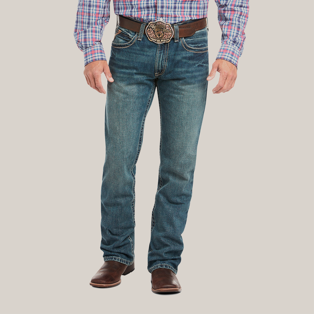 Someone is wearing the M4 Low Rise Boundary Boot Cut Jean in blue, paired with a brown belt featuring an ornate buckle and brown leather shoes. Their plaid shirt is neatly tucked in, highlighting the classic boot cut style. Only the lower half of their body is visible.