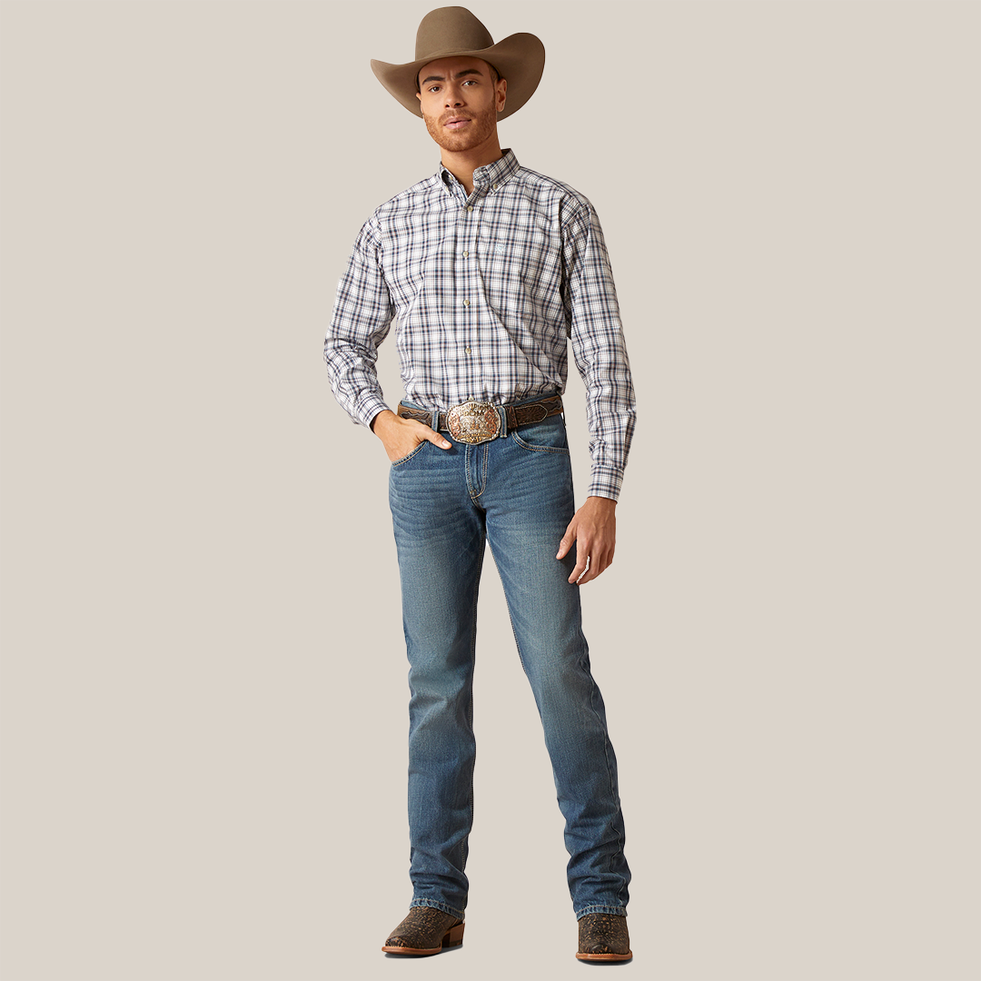A modern cowboy exudes confidence in his M7 Slim Ezra Straight Jeans - Brighton (10045388), paired with a plaid shirt, boots, and classic cowboy hat. His large belt buckle gleams subtly against the plain white background as he casually rests a hand in his pocket.