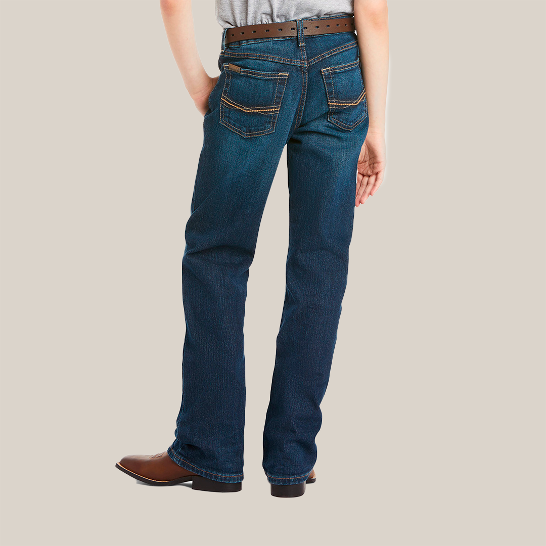 Rear view of a person in Ariat Boys B4 Legacy Dark Wash Relaxed Fit Boot Cut Jeans with visible stitching, paired with a gray shirt, brown belt, and cowboy boots. Their left hand rests in the back pocket.