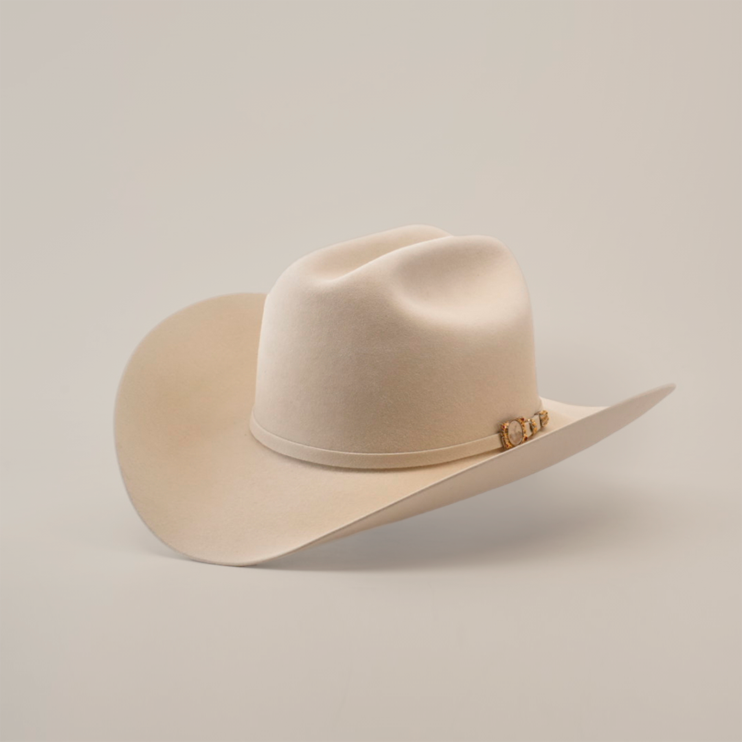 The 100X Independencia Belly, a luxury beige cowboy hat made from premium full beaver fur, boasts a curved brim and decorative band, elegantly showcased on a white surface.