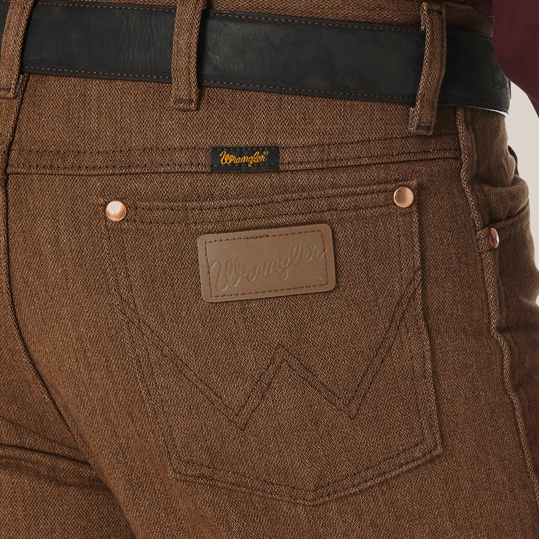 Close-up of a person wearing Wrangler Cowboy Cut Slim Fit Jeans - 100936BKW with a black belt. The jeans feature a branded leather patch above the right pocket and a small tag on the waistband, paired with a dark red shirt for classic style and rugged elegance.