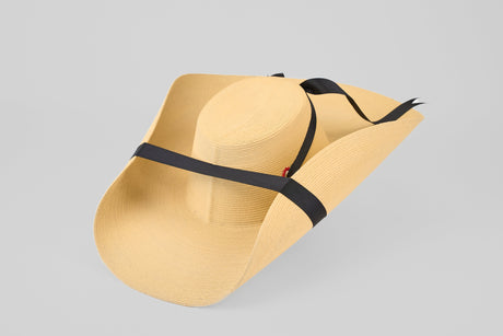 A wide-brimmed, light tan straw hat with a black ribbon wrapped around the crown and chin straps, set against a plain light gray background.