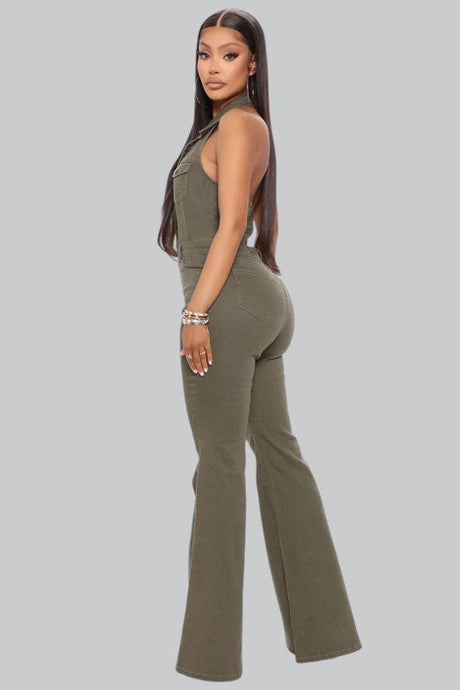 Woman with long, straight hair wearing an olive green sleeveless jumpsuit and bracelets, standing sideways and looking over her shoulder against a plain gray background.