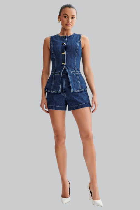 A woman stands against a gray background wearing a sleeveless dark denim button-up top with matching shorts and white heels.