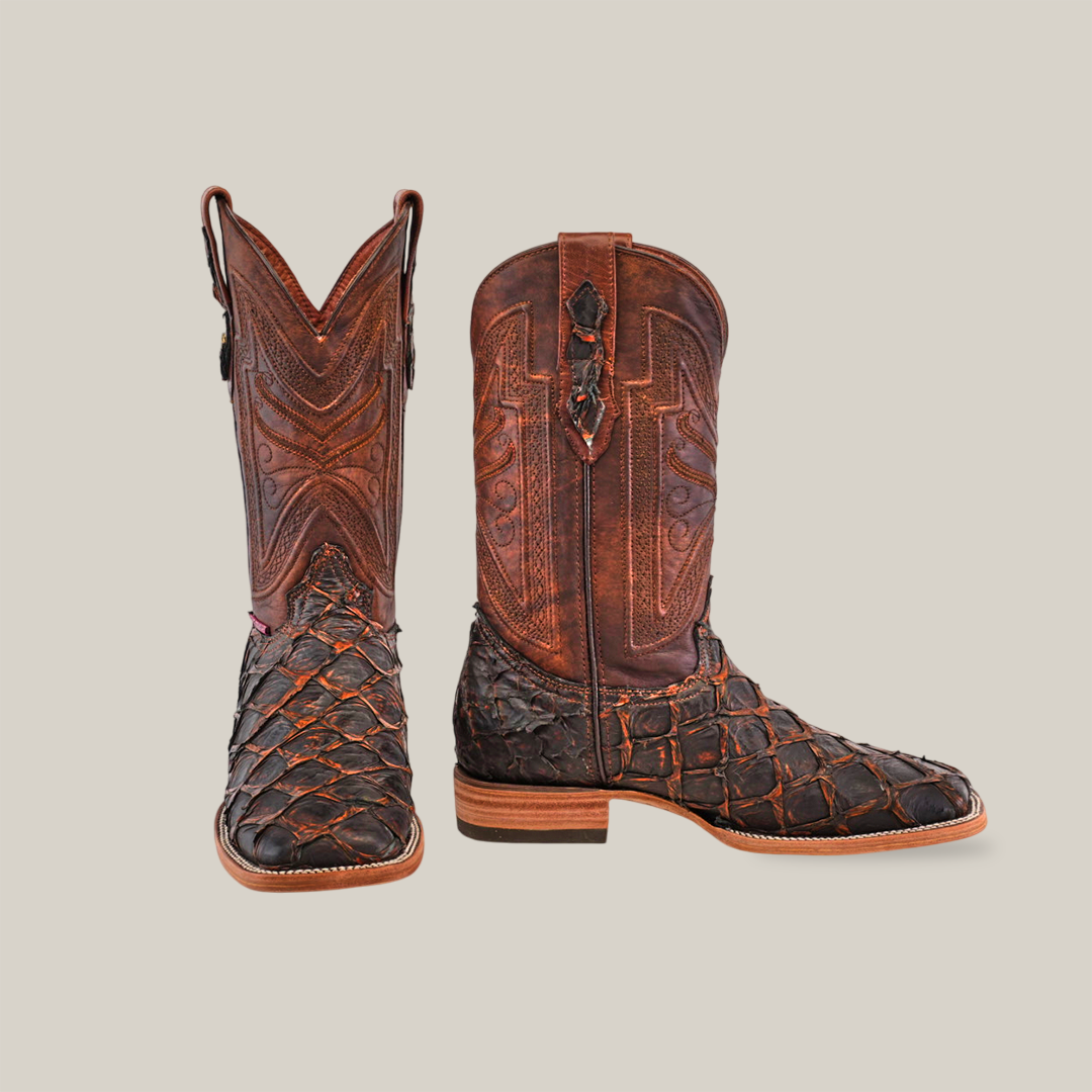 A pair of brown cowboy boots with intricate stitching and a textured, scale-like pattern on the foot and toe, displayed upright on a light beige background.