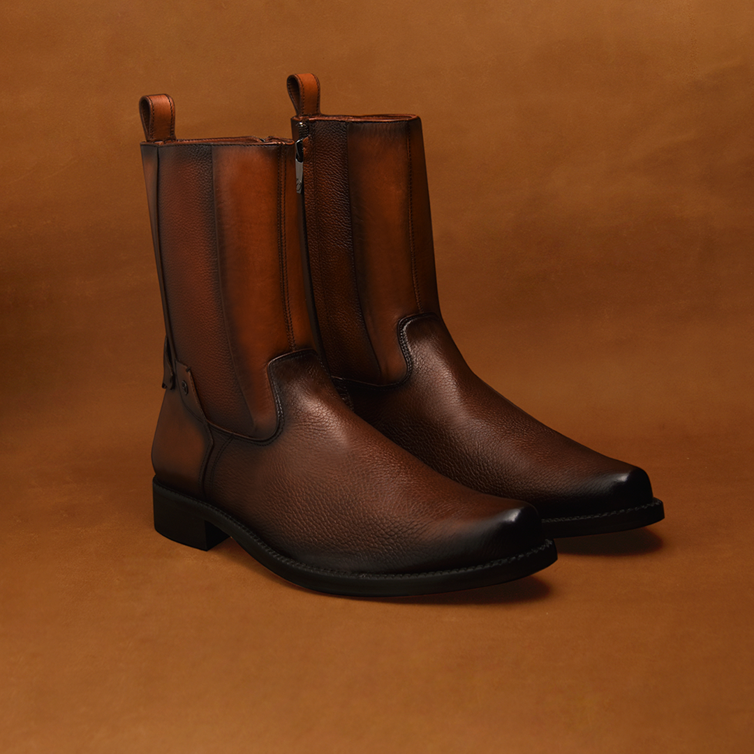 The Honey Deer Zipper - Round Toe is a pair of brown leather boots featuring intricate embroidery and a side zipper, set against a matching brown background. They boast smooth finishes, rounded toes, and small loops at the top for easy wearing.