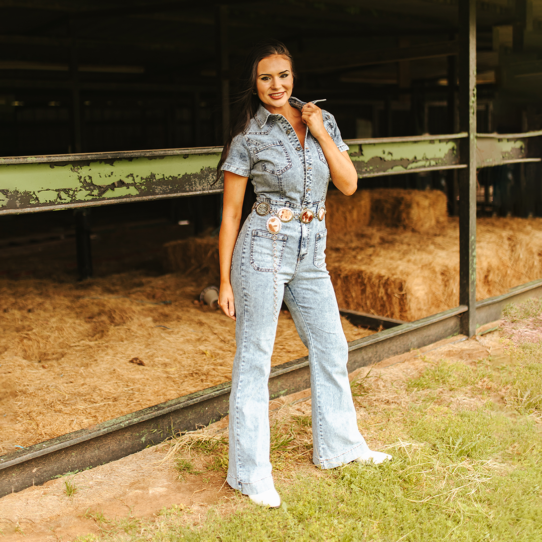 In front of a stable, a woman exudes confidence in the LB Light Denim Jumper JP208-SW. The jumpsuits figure-enhancing silhouette and detailed belt accentuate her style, while her long dark hair and white sneakers add a touch of charm to the rustic backdrop complete with hay bales.