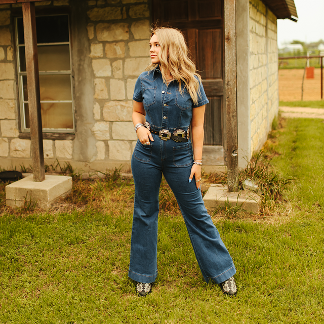 A woman confidently stands on grass near a stone house with wooden accents, wearing the LB Dark Denim Jumper JP208-DW with front button closures. Her long, wavy hair complements the outfits figure-enhancing silhouette and boots, enhancing the rustic outdoor vibe.