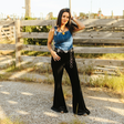 A woman poses outdoors on a gravel path, wearing the LB Denim Texas Buckle Crop, featuring adjustable straps and paired with black flared pants. Shes touching her hair with one hand, set against wooden fencing and grassy surroundings.