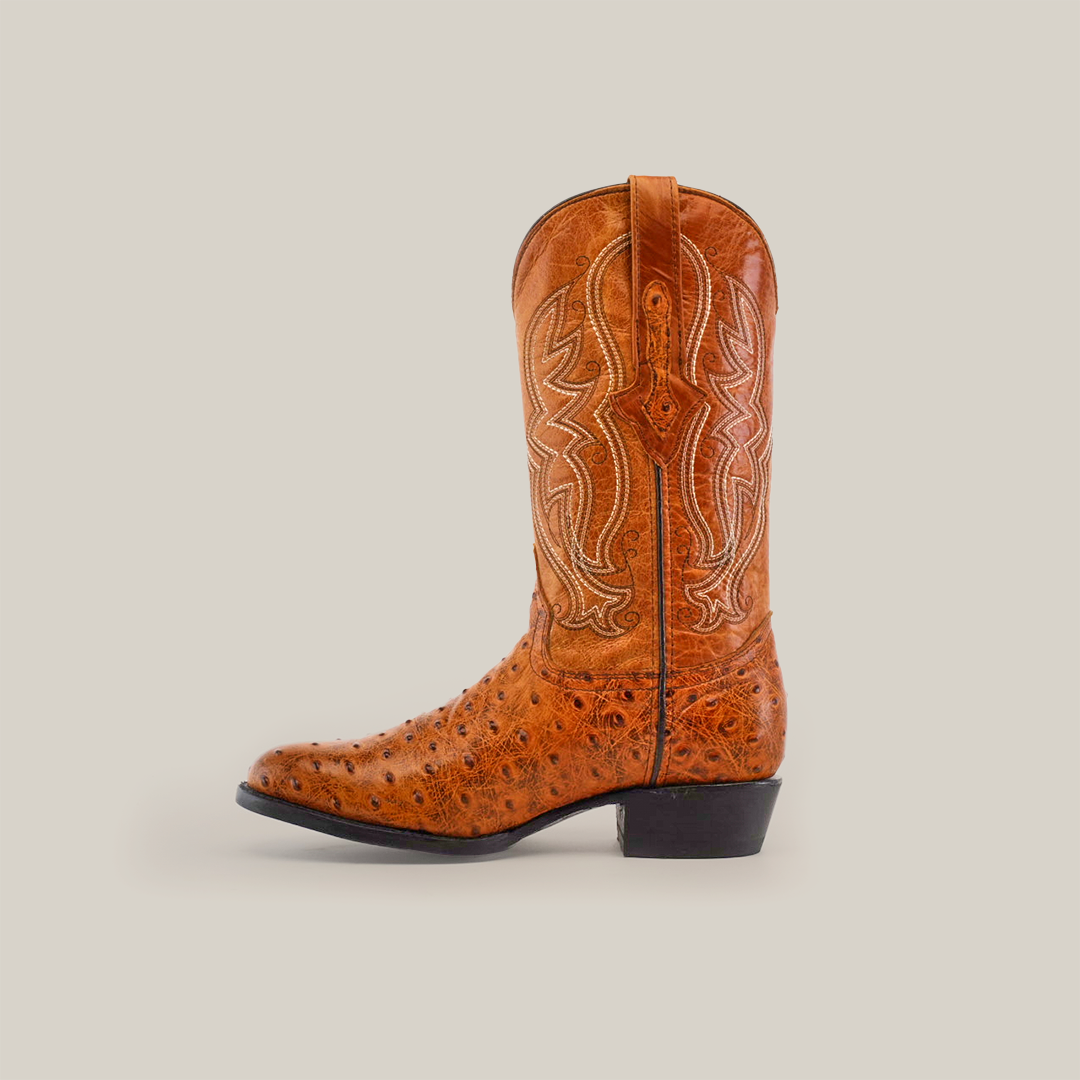 The Karoo Ostrich Print Buttercup boot showcases a round toe with decorative stitching, embossed floral patterns, and an ostrich print texture. It has a leather sole, low heel, and side pull-tabs. Elegantly displayed against a plain white background.