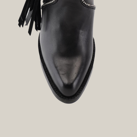 Close-up of the Praga Fringe Tall Shaft Black - J Toe from the Platinum Collection, featuring a polished black leather shoe with a pointed toe and decorative black tassels. White stitching along the edges contrasts with the sleek design on a light background.