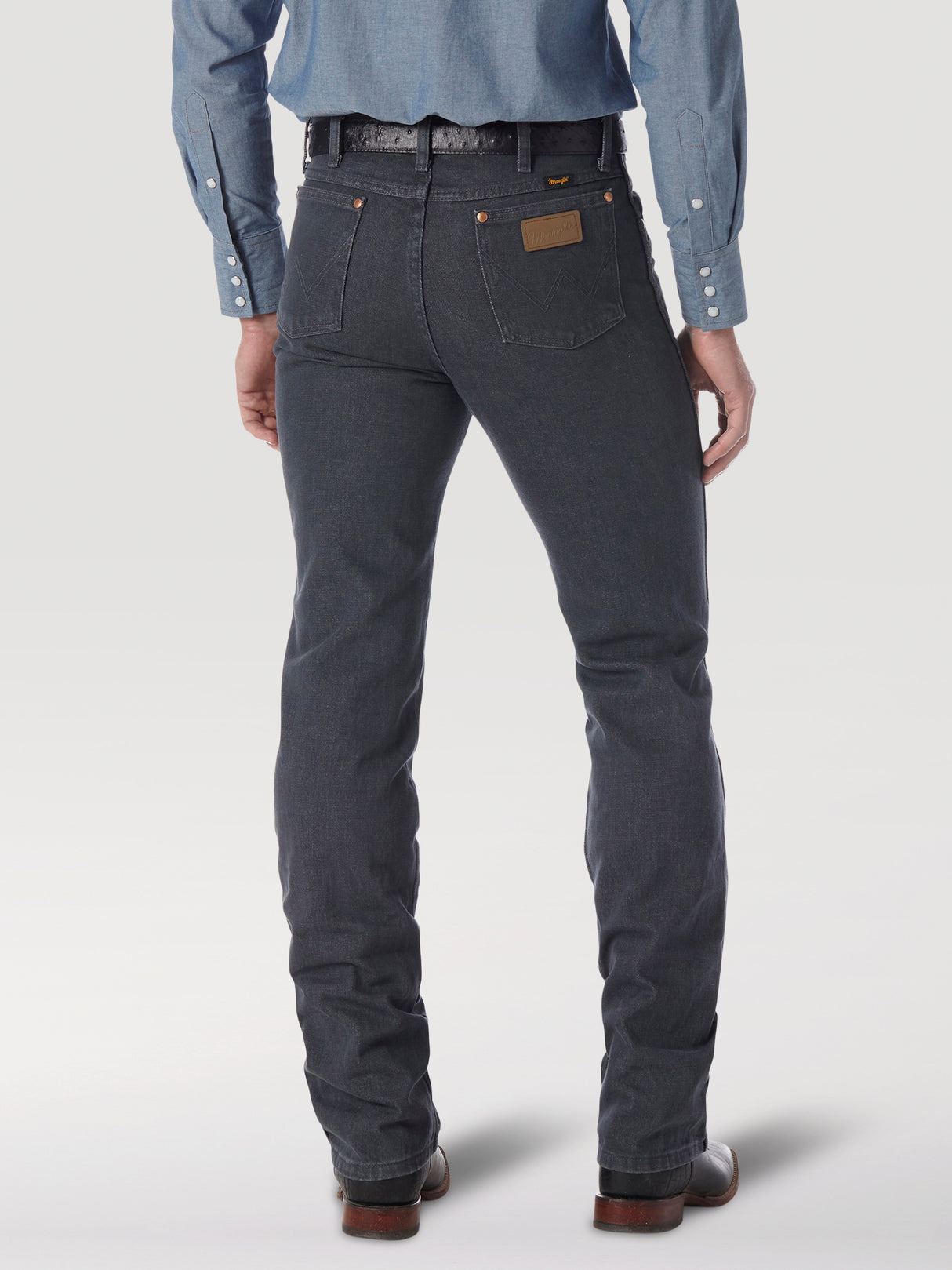 Rear view of a person in dark gray Wrangler Cowboy Cut Slim Fit Jeans - 0936CHG, a blue denim shirt with rolled-up cuffs, and brown shoes. A leather patch graces the back pocket. Set against a plain light background, the outfit exudes timeless appeal.