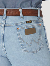 Close-up of a person in Wrangler Cowboy Cut Slim Fit Jeans - 0936GBH, light blue with a brown leather belt. The premium denim displays the classic W stitching on the back pocket with a leather Wrangler patch above it.