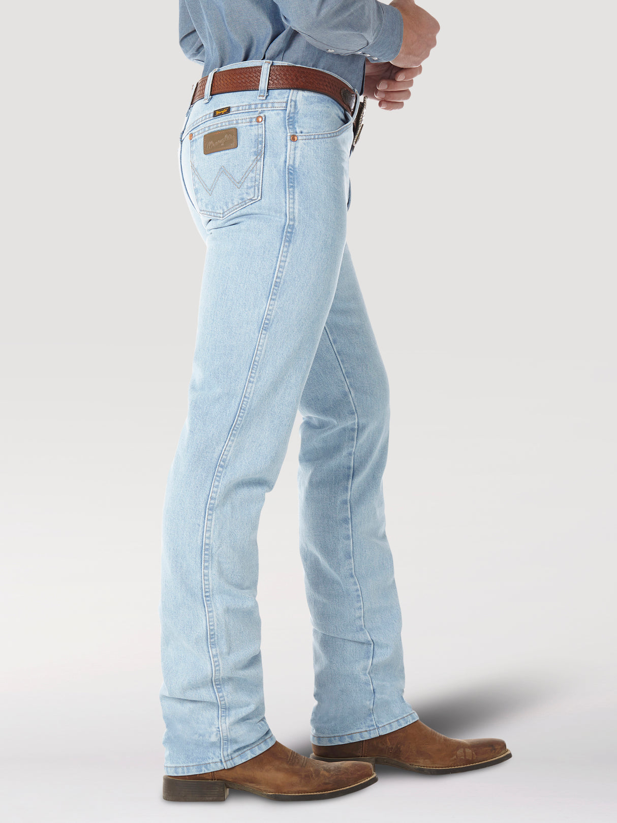 A side view shows a person in light blue Wrangler Cowboy Cut Slim Fit Jeans - 0936GBH, with a brown belt and cowboy boots, standing against a plain background. The jeans feature classic stitching on the back pocket.