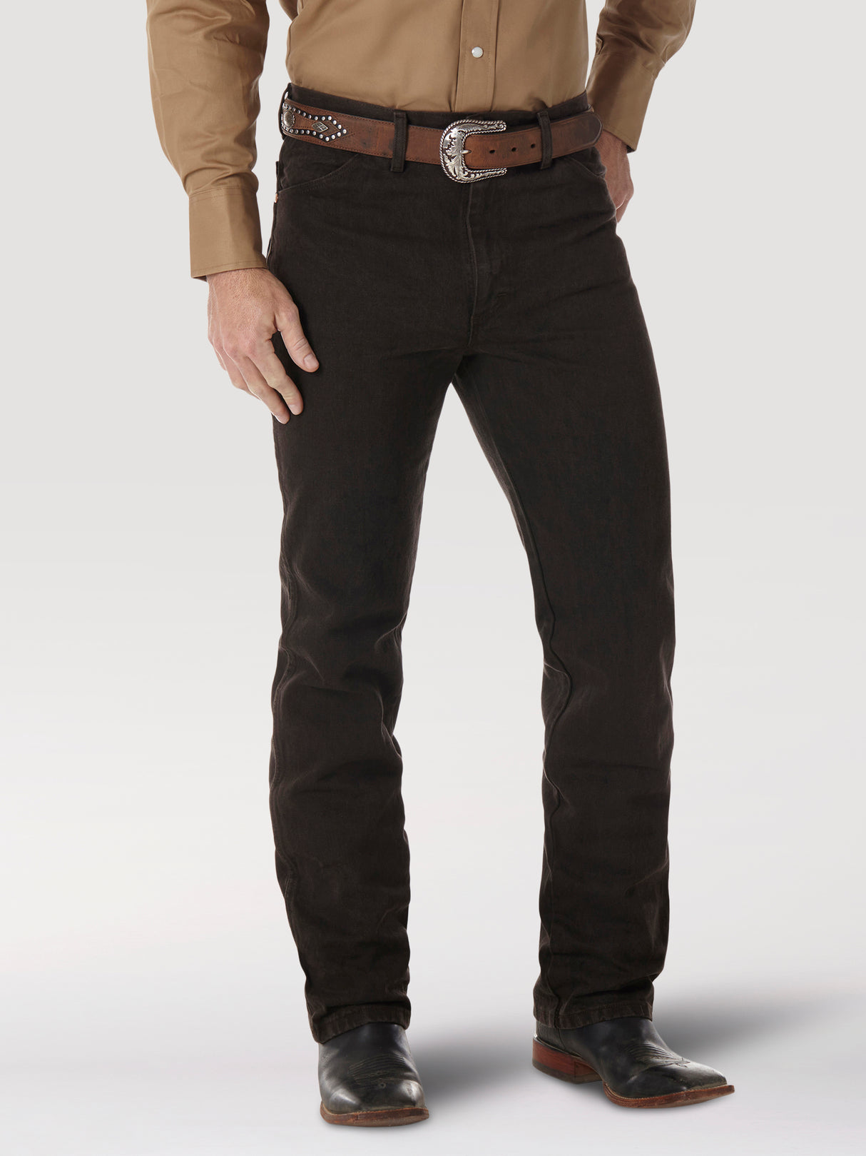 A person wearing a tan long-sleeve shirt, Wrangler Cowboy Cut Slim Fit Jean (0936KCL), a studded brown belt with a large buckle, and black boots. The backdrop is plain white.