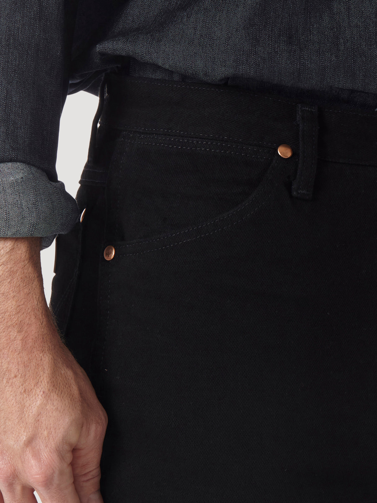 A person in a dark denim shirt with rolled sleeves and wearing Wrangler Cowboy Cut Slim Fit Jean - 0936WBK. The photo highlights the premium denims pocket, showing stitching and copper rivets, as their hand rests relaxed by their side.