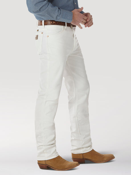 Wearing a blue shirt tucked into Wrangler Cowboy Cut Slim Fit Jeans in white (0936WHI), accessorized with a patterned belt and brown cowboy boots, the person is shown from the waist down against a plain background, highlighting the essence of Cowboy Cut style.