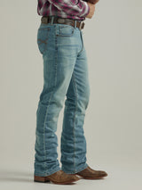 A person in Wrangler 20X Mens Light Wash Shade Bootcut Stretch Jeans - 112335407, brown leather cowboy boots, a brown belt, and a red plaid shirt stands sideways with hands clasped at their waist against a neutral background.