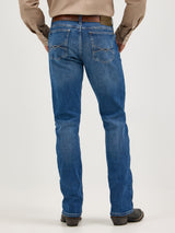 A person in bootcut jeans wears a beige long-sleeve shirt, seen from the back. The Wrangler 20X No. 42 features premium denim with a classic design and a Wrangler patch on the pocket. Black boots and a brown belt complete the look against a plain, light background.