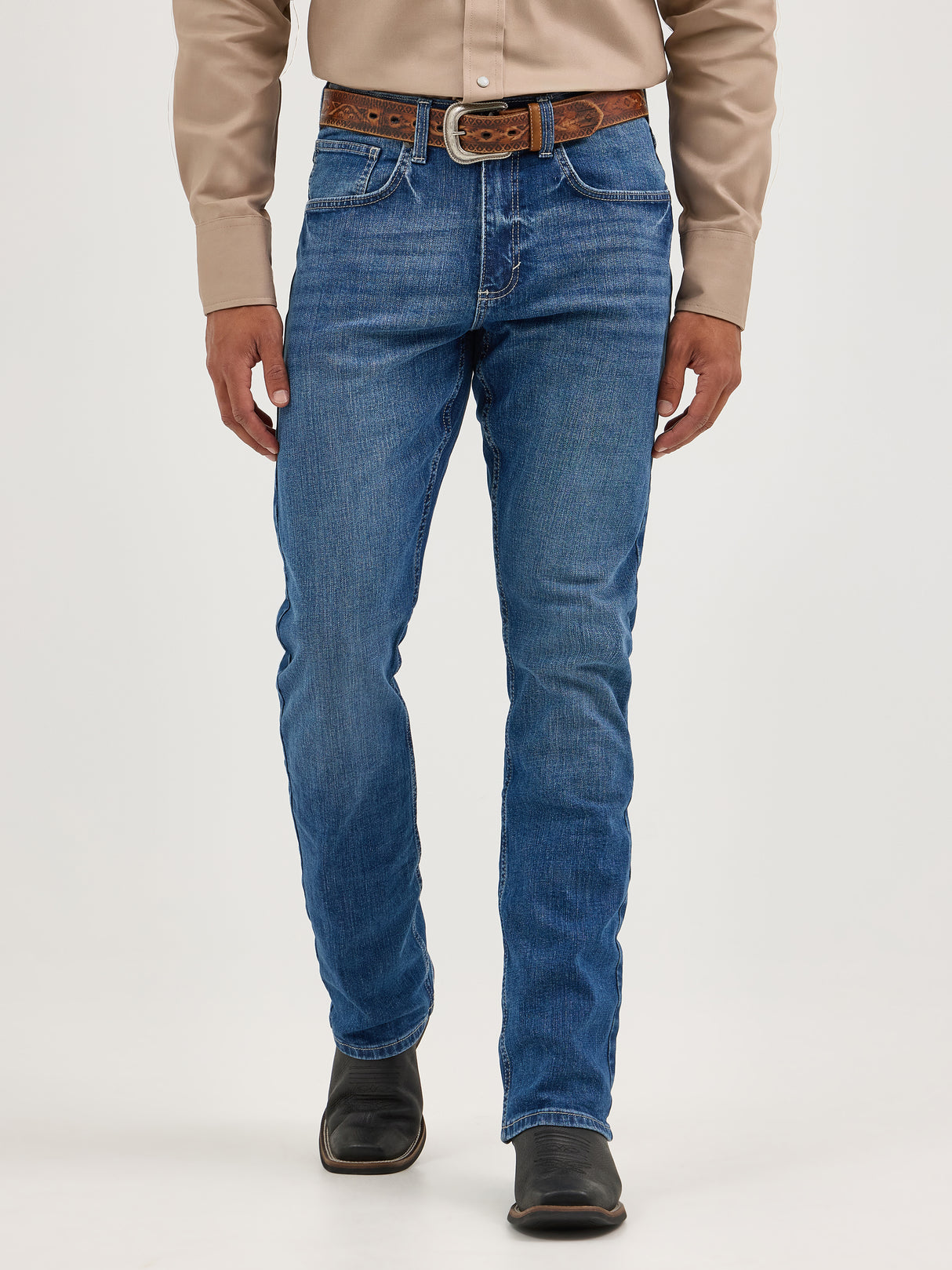 A person in a beige long-sleeve shirt, blue jeans, and a brown belt stands before a plain background. They wear Mens Wrangler 20X No. 42 Vintage Bootcut Jeans with the iconic patch, hands at their sides, while black boots complete the outfit.