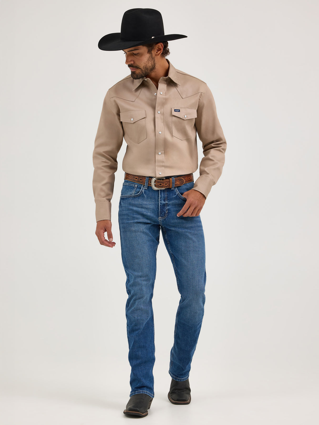 A man in a cowboy hat stands against a plain white background, wearing a beige long-sleeved shirt and Mens Wrangler 20X No. 42 Vintage Bootcut Jeans crafted from premium denim. Dark boots complete his rugged look as he gazes downward, embodying timeless Western style.