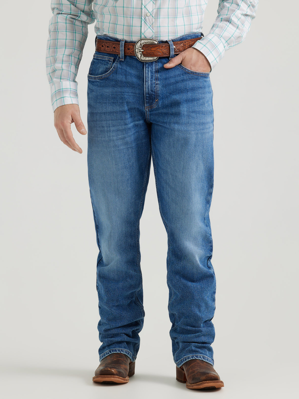 A person wearing Wrangler 20x Mens Medium Wash Backwater Vintage Bootcut Stretch Denim Jeans with a brown tooled leather belt, plaid shirt, and brown cowboy boots. The image highlights the lower half of the body against a plain background.