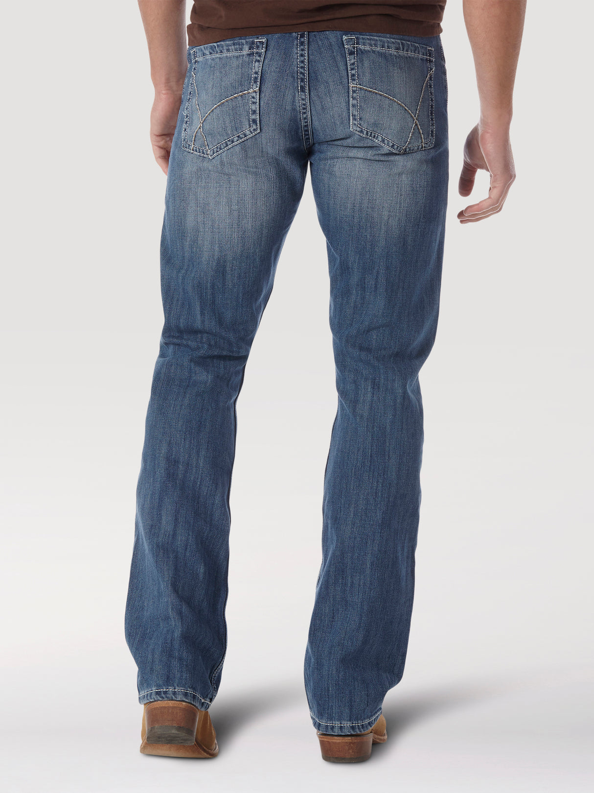 Back view of a person wearing Wrangler 20X No. 42 Vintage Bootcut Jeans (1042MWXLB) and a brown shirt, standing against a plain backdrop. The jeans have decorative stitching on the back pockets and are paired well with brown shoes.