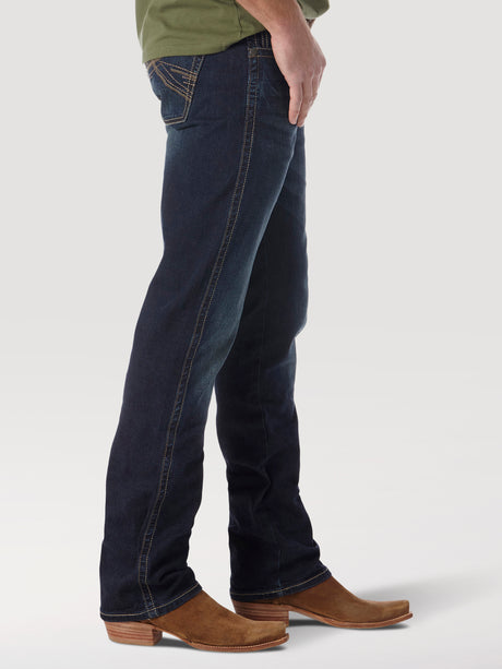 Side view of a person wearing Mens Wrangler 20X No. 42 Vintage Bootcut Jeans (42MWXRD) in dark blue with light brown cowboy boots, against a white background. The jeans, featuring premium contrast stitching on the back pocket, are visible from the chest down.