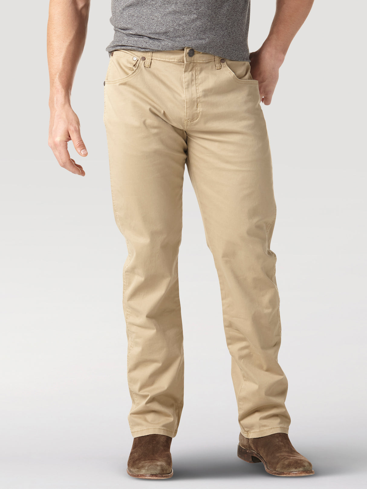 A person wearing a gray t-shirt, beige pants, and brown suede shoes stands against a neutral background. Their left hand is by their side, and their right hand rests casually in the pocket of their Mens Wrangler Retro Slim Fit Straight Leg Pant - 1088MWZFN.