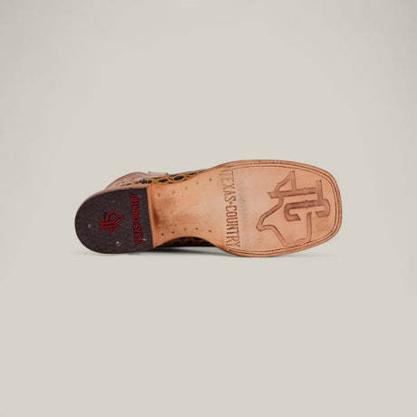 The Jonh - Orix - Square Toe boot features a flat tan leather sole stamped with “TEXAS COUNTRY” and a bold TC logo. Its dark heel displays a red Texas Country emblem set against a plain, light background.
