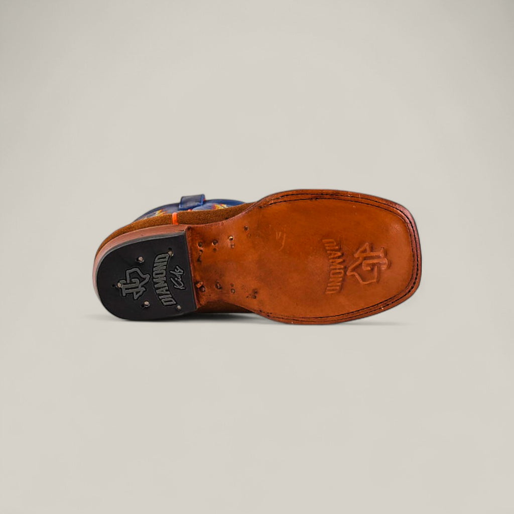 A close-up of the Diamond Suede Ocre - Square Toe - Kids shoe sole shows brown leather with stitched edges, a black rubber heel cap, and embossed logos on both the sole and heel. The background is plain and light-colored.
