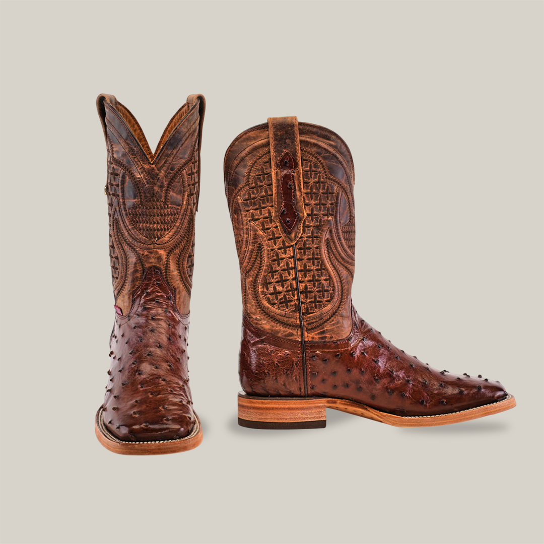 Two brown Exotic Full Quilt Ostrich cowboy boots with intricate patterns, square toes, and sturdy heels are displayed against a light background. One boot stands upright while the other is turned slightly to showcase its design.