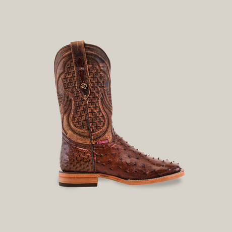 The Exotic Full Quilt Ostrich - Brown - Square Toe boot features intricate stitching, a textured ostrich leather design, a red branded tag near the ankle, pull strap, and two-tone wooden heel. Its displayed stylishly on a neutral backdrop.