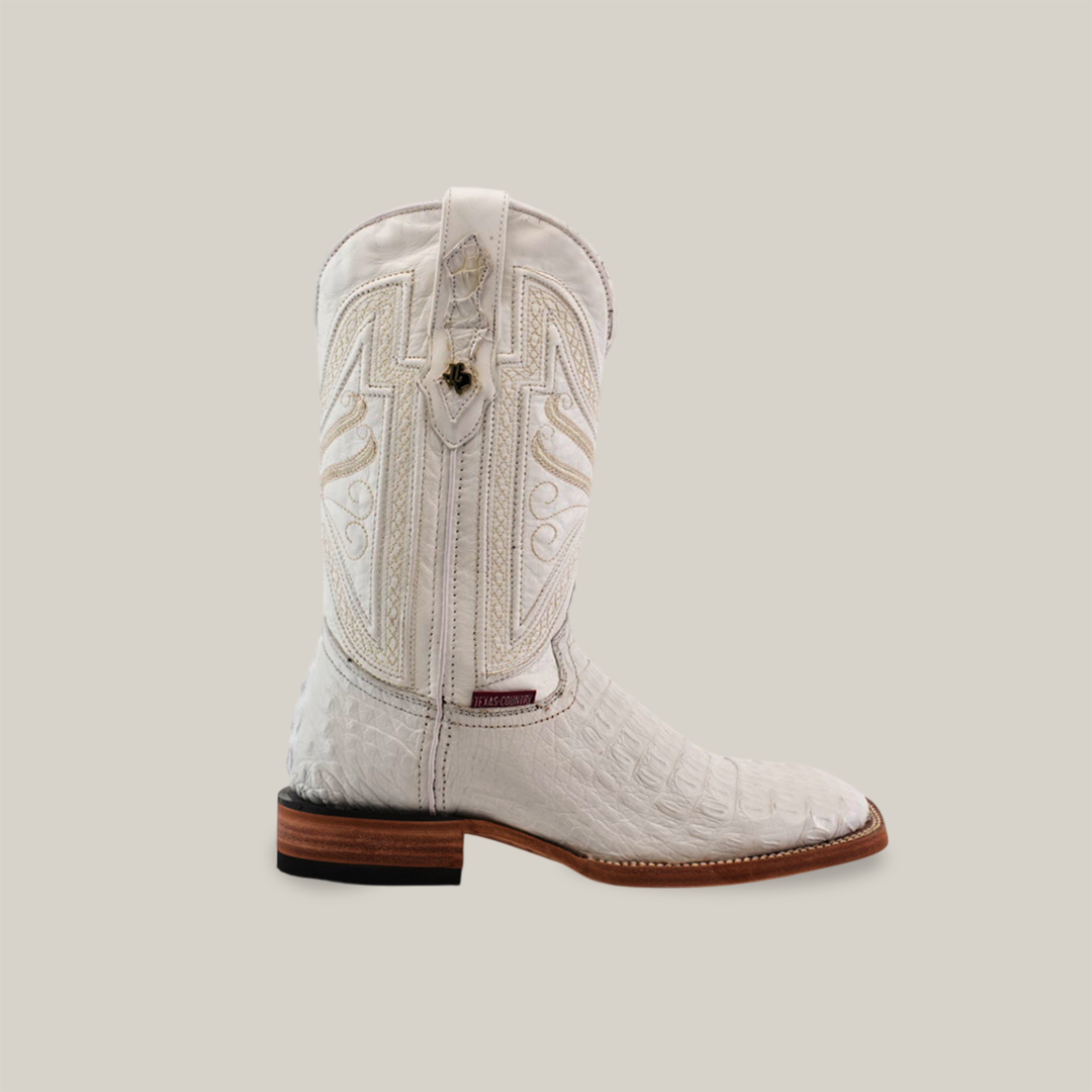 The Exotic Caiman Horn Back boot in white showcases quality craftsmanship with intricate stitching and embossed patterns. It features a square toe and a brown wooden heel, all set against an off-white backdrop.