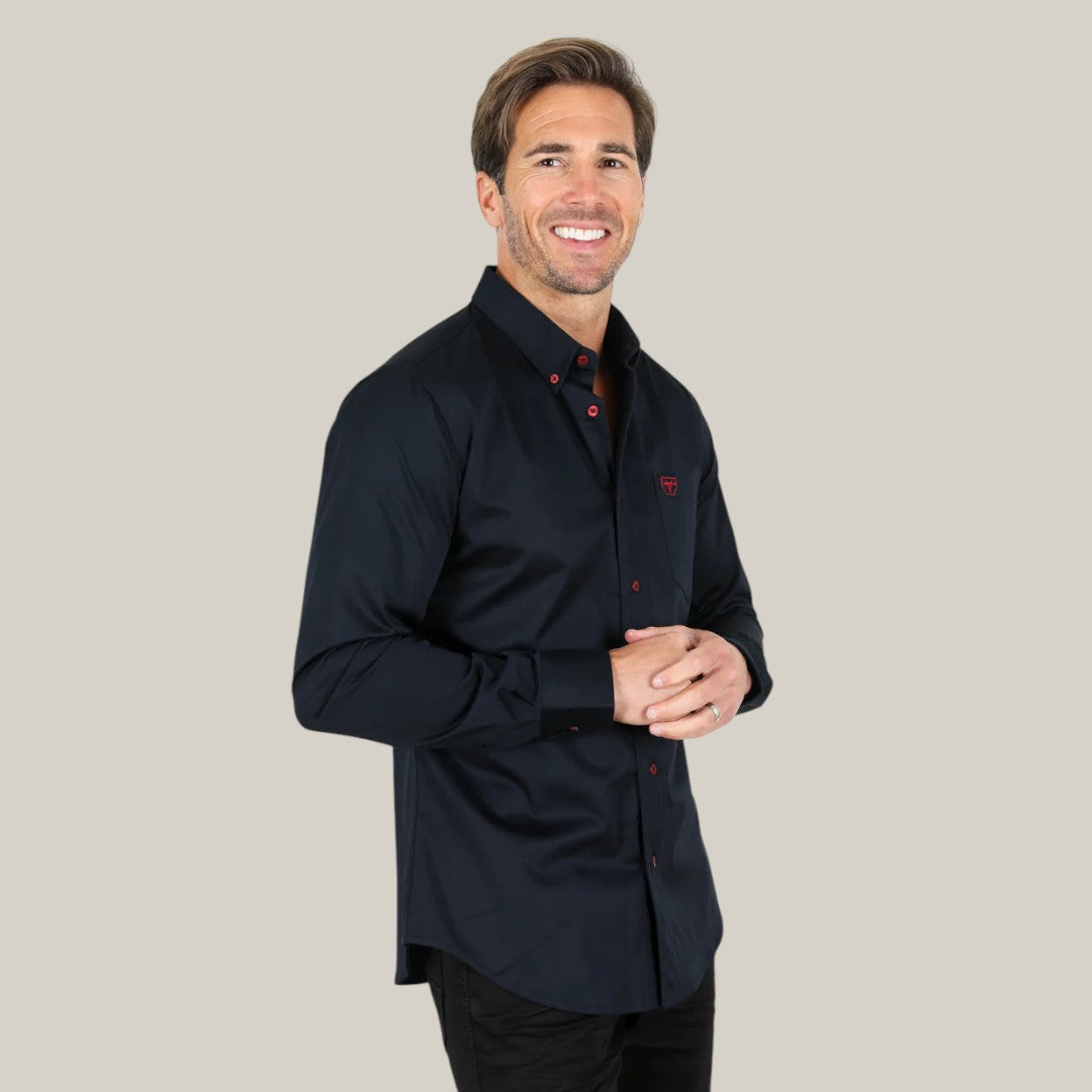 A smiling man wearing the Men’s Single Pocket Logo Modern Fit Stretch Dress Shirt in navy, featuring striking red buttons, stands against a beige background. The stretch fabric ensures comfort as he clasps his hands in front.